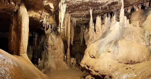 A dimly lit cave interior featuring stalactites and stalagmites, with wet, rocky surfaces and a natural, rugged landscape.