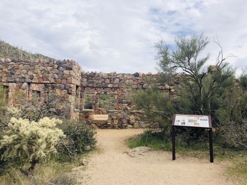 Ruins of a stone building surrounded by desert vegetation and a sign, under a cloudy sky.