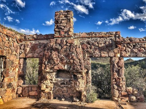 Ruins of a stone building with open windows, set against a blue sky with scattered clouds and desert vegetation.