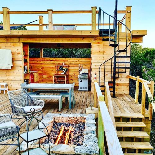 A cozy wooden deck with a spiral staircase, outdoor kitchen, seating area, and a fire pit surrounded by stones.
