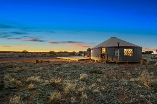 A cozy yurt at sunset, surrounded by open land and a clear blue sky, with warm light glowing from the windows.
