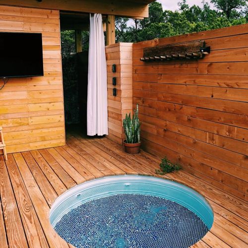 A wooden deck features a circular hot tub, a potted plant, and a TV mounted on the wall, surrounded by natural greenery.