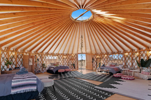 Interior of a spacious yurt with wooden beams, two beds, rugs, and a skylight, surrounded by desert scenery.