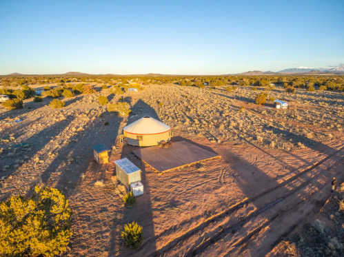 Aerial view of a yurt in a vast desert landscape during sunset, surrounded by sparse vegetation and distant mountains.
