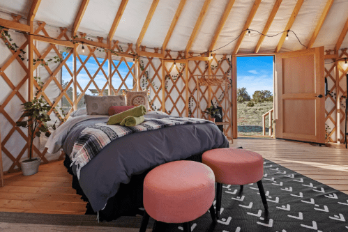 Cozy yurt interior with a bed, pink stools, and a view of the outdoors through an open door.