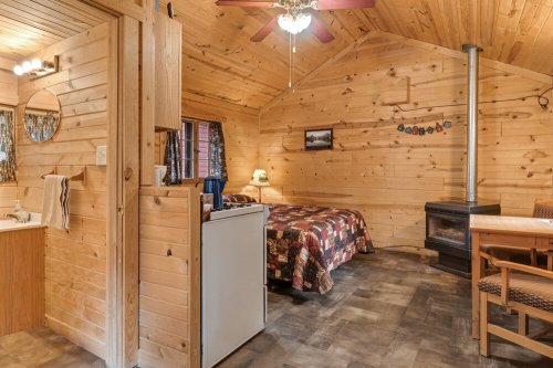 Cozy wooden cabin interior featuring a bed, kitchenette, dining table, and a wood stove. Warm, rustic decor.