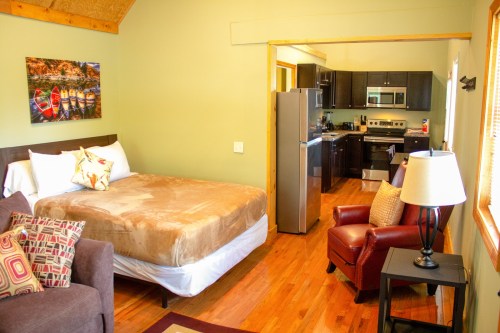Cozy room with a bed, armchair, and kitchenette featuring modern appliances and wooden floors.