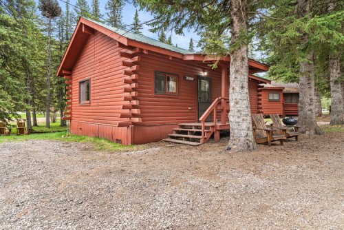 A rustic red log cabin surrounded by trees, with a gravel path and wooden chairs in front.