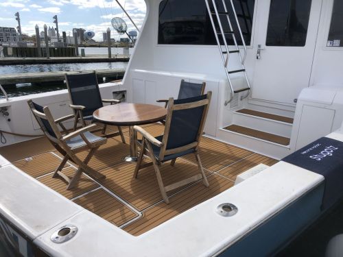 A boat deck featuring a round table and four wooden chairs, with a staircase and city skyline in the background.