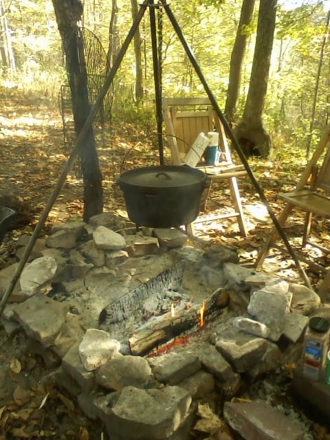 A campfire setup with a pot hanging over stones, surrounded by autumn leaves and a wooden chair nearby.