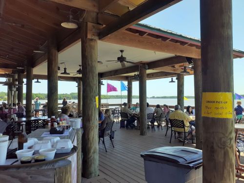 A waterfront dining area with people seated at tables, enjoying a meal under a wooden pavilion. Colorful flags flutter nearby.
