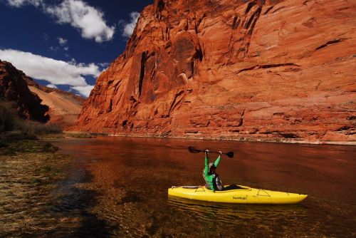 A person in a green kayak performs a handstand on a calm river, surrounded by red rock cliffs and blue skies.