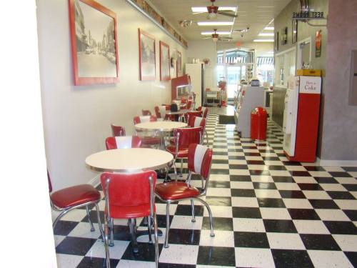 Retro diner interior with red and white decor, checkered floor, and round tables. Vintage soda fountain in the background.
