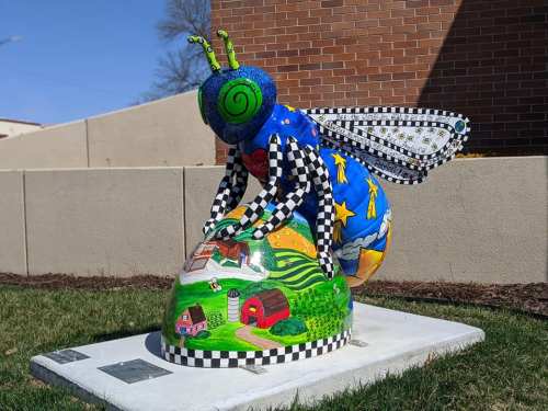Colorful bee sculpture with a whimsical design, kneeling over a painted landscape, set against a brick wall.