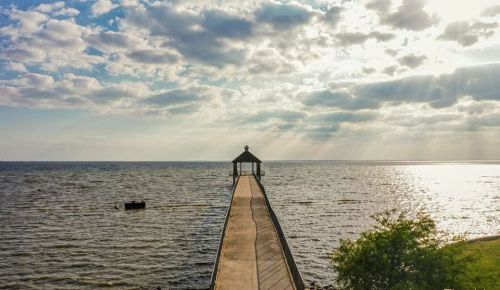 A serene pier extends into a calm body of water under a cloudy sky, with sunlight breaking through the clouds.