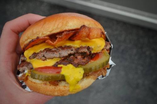 A hand holding a juicy burger with bacon, cheese, pickles, ketchup, and mustard on a toasted bun.