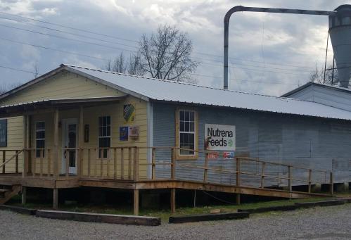 A small, yellow and gray building with a porch, featuring a sign for Nutrena Feeds and a grain silo in the background.