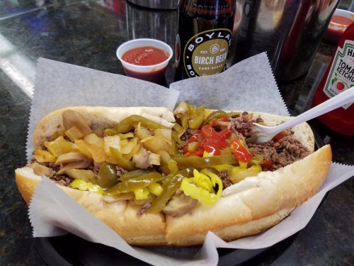 A loaded sandwich with beef, peppers, onions, and mushrooms, served with a bottle of birch beer and a side of sauce.