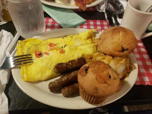 A plate with an omelette, sausage links, hash browns, and two muffins, set on a checkered tablecloth.