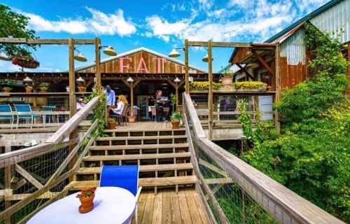 A rustic restaurant with wooden steps, outdoor seating, and vibrant greenery under a blue sky.