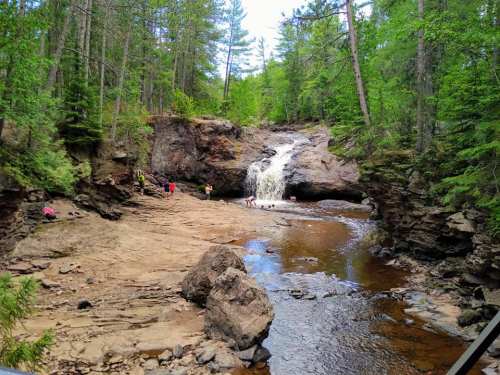 A serene waterfall cascades into a rocky pool, surrounded by lush green trees and people enjoying the natural setting.