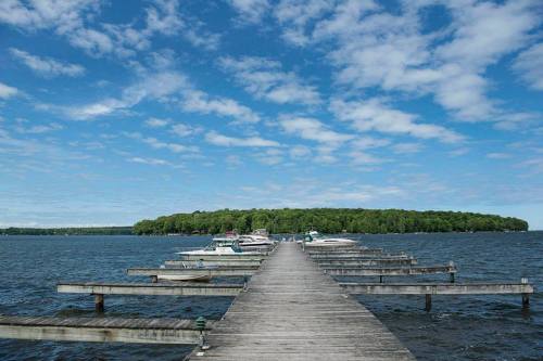 A wooden dock extends into a calm lake, lined with boats, under a blue sky with scattered clouds.