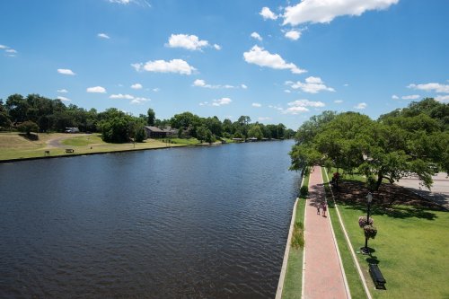 A serene river scene with a walking path, lush greenery, and blue skies dotted with clouds.