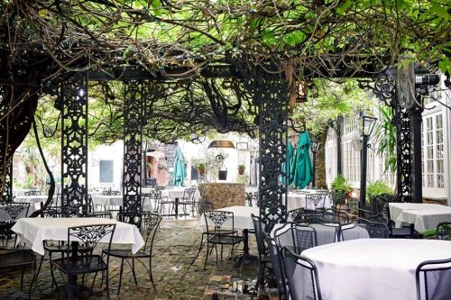 A charming outdoor restaurant with wrought iron tables and chairs, surrounded by lush greenery and a canopy of vines.