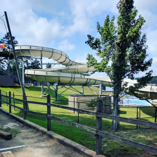 A water slide twists down into a pool area, surrounded by grass and trees, with a wooden fence in the foreground.