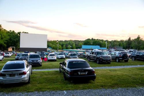 A drive-in movie theater with numerous cars parked in front of a large outdoor screen at sunset.