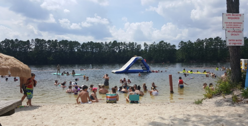 A busy beach scene with people swimming and playing on an inflatable slide, surrounded by trees and a sandy shore.