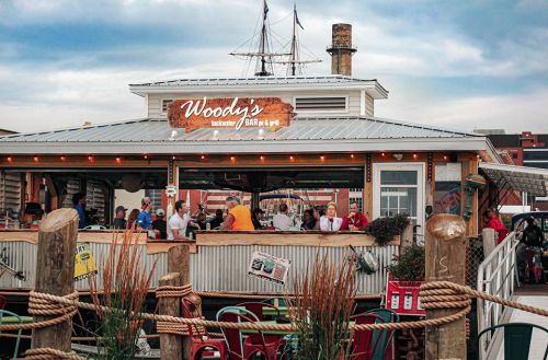 A lively waterfront restaurant, Woody's, with outdoor seating and a ship's mast in the background under a cloudy sky.