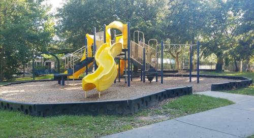Colorful playground with yellow slides and climbing structures, surrounded by trees and a gravel surface.
