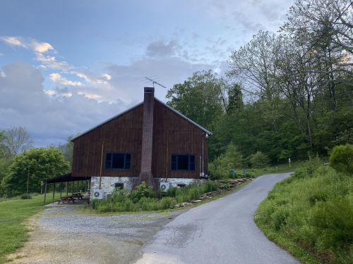 A rustic wooden house with a stone foundation, surrounded by greenery, sits at the end of a winding gravel road.
