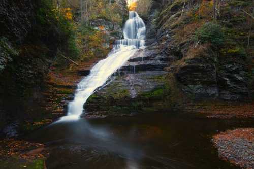 A serene waterfall cascades over rocky cliffs into a tranquil pool, surrounded by autumn foliage.