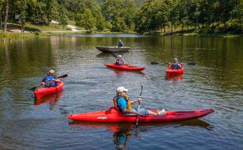 Five people in red kayaks paddle on a calm lake surrounded by trees and a small boat in the background.