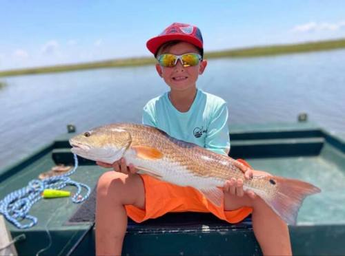 A young boy in sunglasses and a cap proudly holds a large fish while sitting on a boat in a sunny outdoor setting.