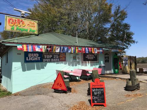 A colorful dive bar with a green roof, outdoor seating, and signs for events and drinks, set by a waterway.