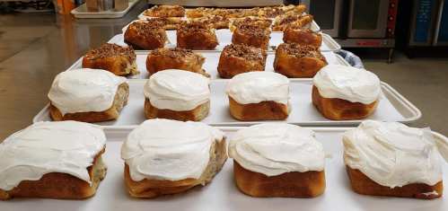 Freshly baked cinnamon rolls with cream cheese frosting and pecan-topped pastries on trays in a kitchen.