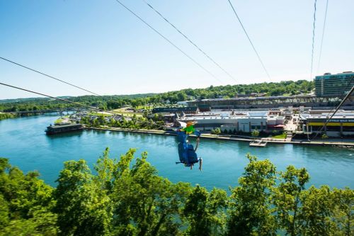 A person zip-lining over a river with a scenic view of trees and buildings along the waterfront.