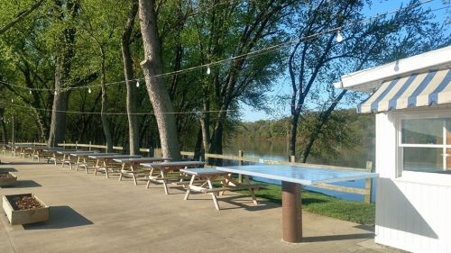 Outdoor seating area by a river, featuring picnic tables and string lights, surrounded by trees on a sunny day.