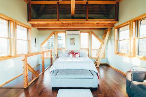 Cozy bedroom with wooden beams, large windows, and a staircase leading to a loft area, featuring a bed and seating.