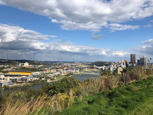 A panoramic view of Pittsburgh, featuring the skyline, rivers, and green hills under a partly cloudy sky.