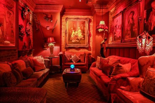 Cozy, dimly lit lounge with red walls, plush sofas, vintage artwork, and a glowing blue orb on a coffee table.