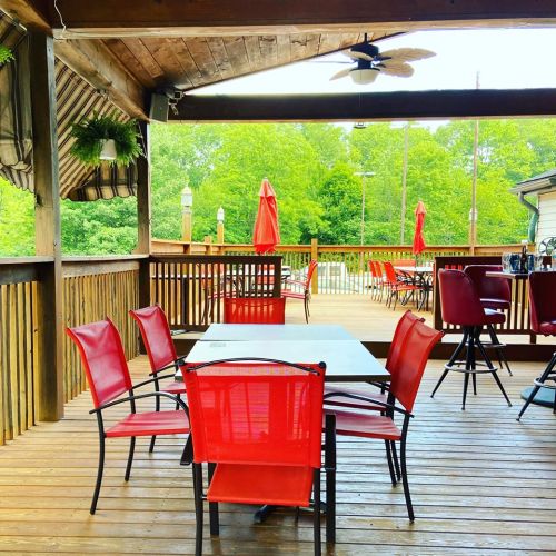 A wooden deck with red chairs and tables, surrounded by greenery and umbrellas, creating a cozy outdoor dining space.