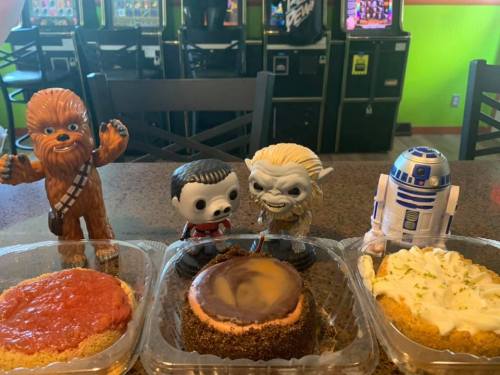 Four figurines stand beside three colorful desserts on a table, with arcade machines visible in the background.