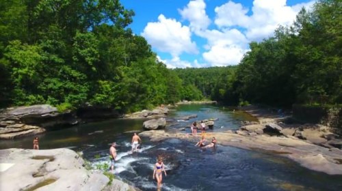 People enjoying a sunny day by a river, swimming and playing among rocks and lush green trees.