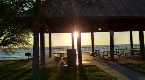 Sunset view through a pavilion by the water, with picnic tables and trees silhouetted against the glowing horizon.