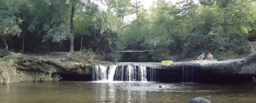 A serene scene of a small waterfall cascading into a calm pool, with two people relaxing nearby on the shore.