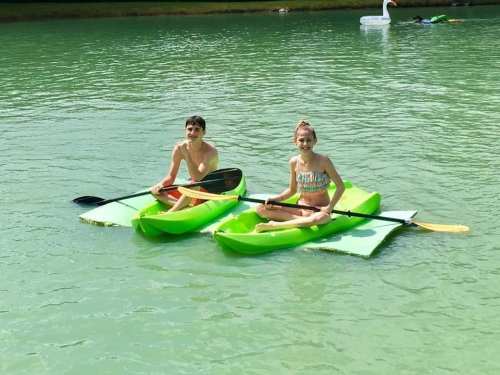 Two children sit on bright green kayaks in a calm, green-tinted lake, enjoying a sunny day.